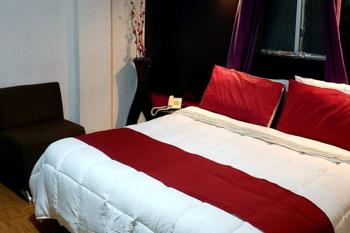 Een bed met rode en witte beddengoed, een nachtkastje met een telefoon, en een stoel in een kamer met een raam en gordijnen.