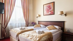A hotel room with two beds, a TV, a painting, and a nightstand with a lamp and bottles. - Conviva Hotel (Panevezys)