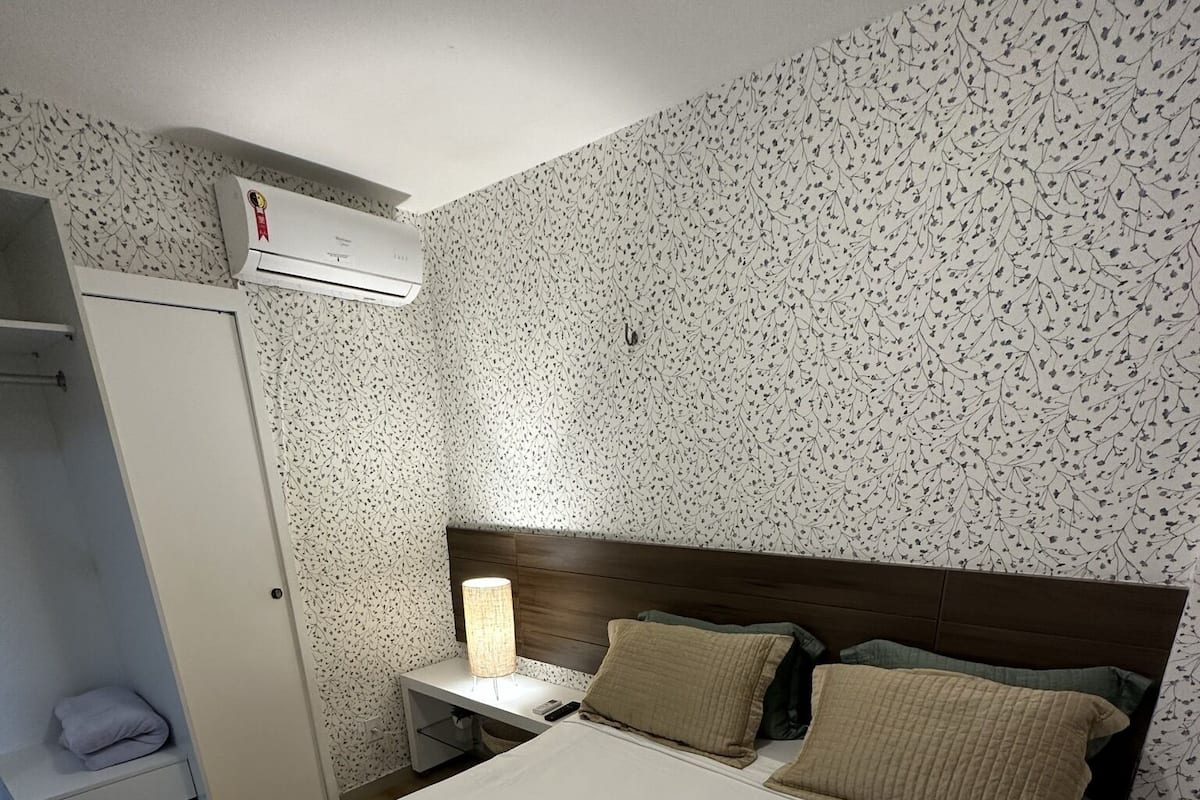 Ein ordentlich bezogenes Bett mit gefaltetem Handtuch, einer Nachttischlampe und einem Nachttisch mit Fernbedienung. Eine Garderobe mit Spiegel ist links im Bild zu sehen.