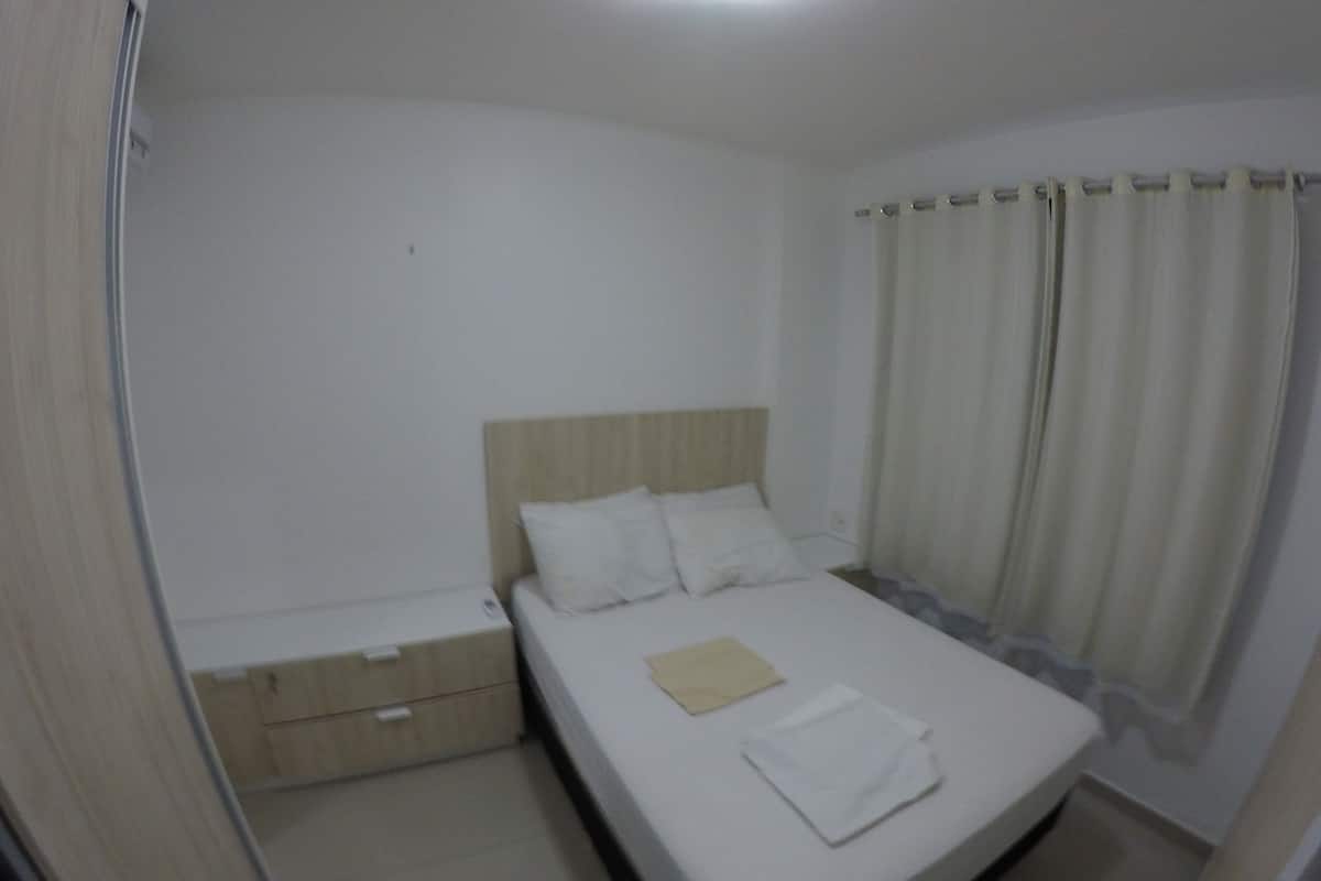 Ein Zimmer mit einem Einzelbett, einem Holzkopfende, weißer Bettwäsche, einem Nachttisch und einem Vorhang.