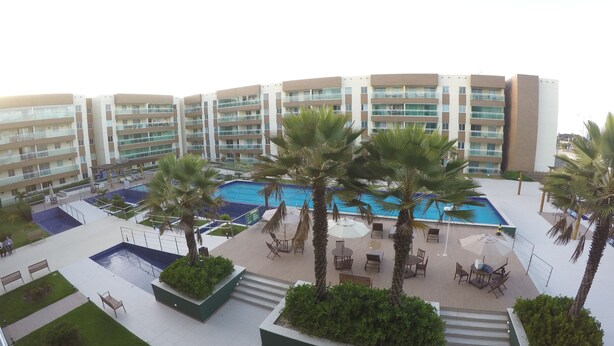A hotel with multiple balconies, a central swimming pool surrounded by palm trees and lounge chairs, and a modern architectural design.