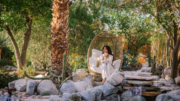 A person sitting in a wicker chair by a small water feature surrounded by rocks and plants.