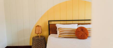 Quarto com cama, mesa de cabeceira, abajur e um banquinho estampado.
