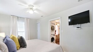 Un dormitorio con cama, ventilador de techo, televisor y lavandería visible a través de una puerta abierta.
