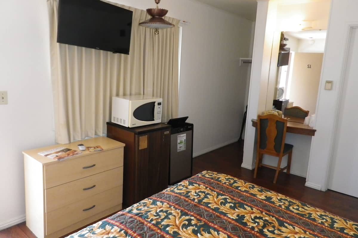 A bedroom with a bed, a dresser, a microwave, a desk with a chair, a mirror, and a television mounted on the wall.