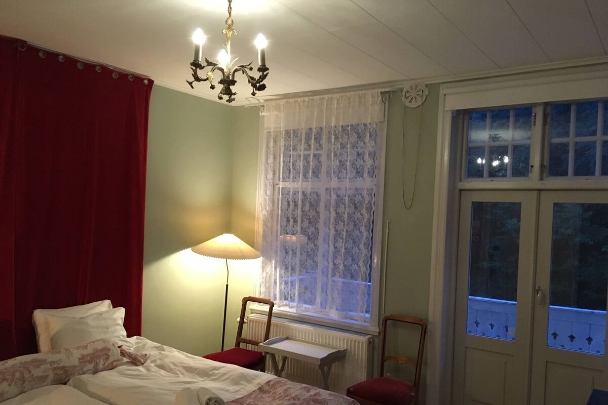 Une chambre à coucher comprenant un lit, un bureau, une chaise, une fenêtre avec des rideaux, un radiateur et un lustre.
