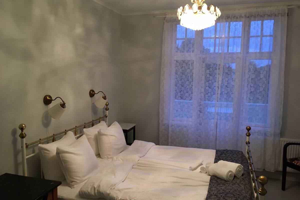 Un lit bien fait, avec du linge de lit blanc, un lustre et une fenêtre ornée de rideaux en dentelle.