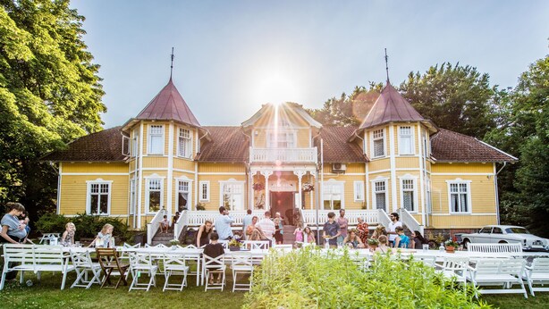 Un grand bâtiment jaune, doté d’une entrée centrale et de deux tours, entouré de tables et de chaises blanches, ce qui laisse supposer qu’un événement ou une réception en plein air y est organisé.