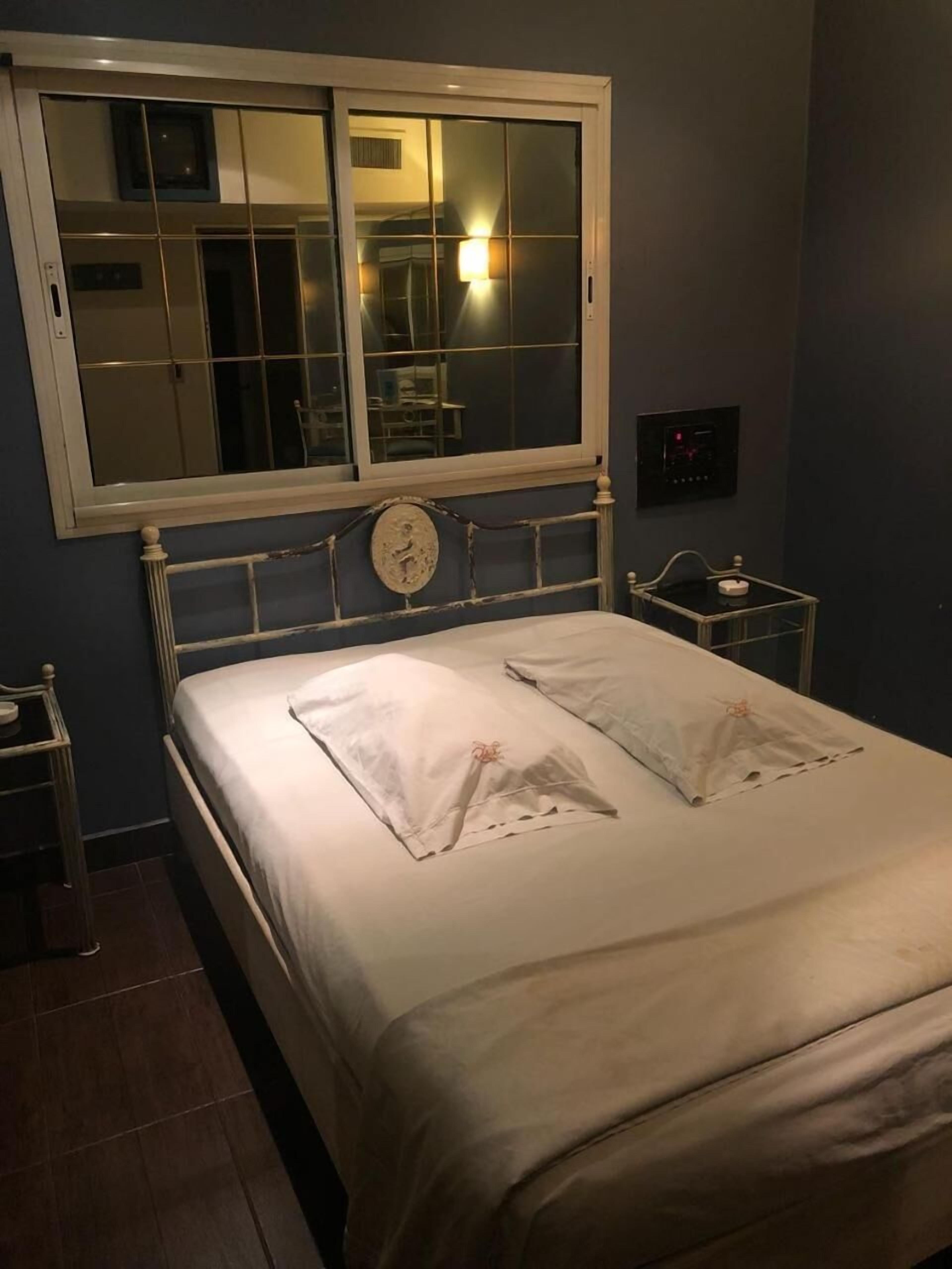 A neatly made bed with white linens, a bedside table with a lamp, and a window with blinds.