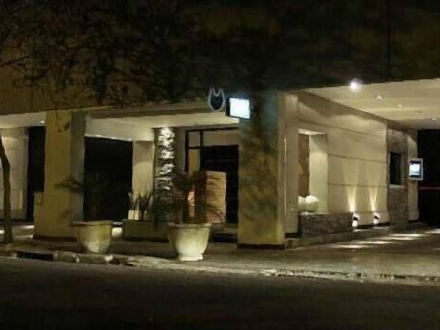 Fachada de hotel iluminada de noche, con plantas en macetas y una señal visible.
