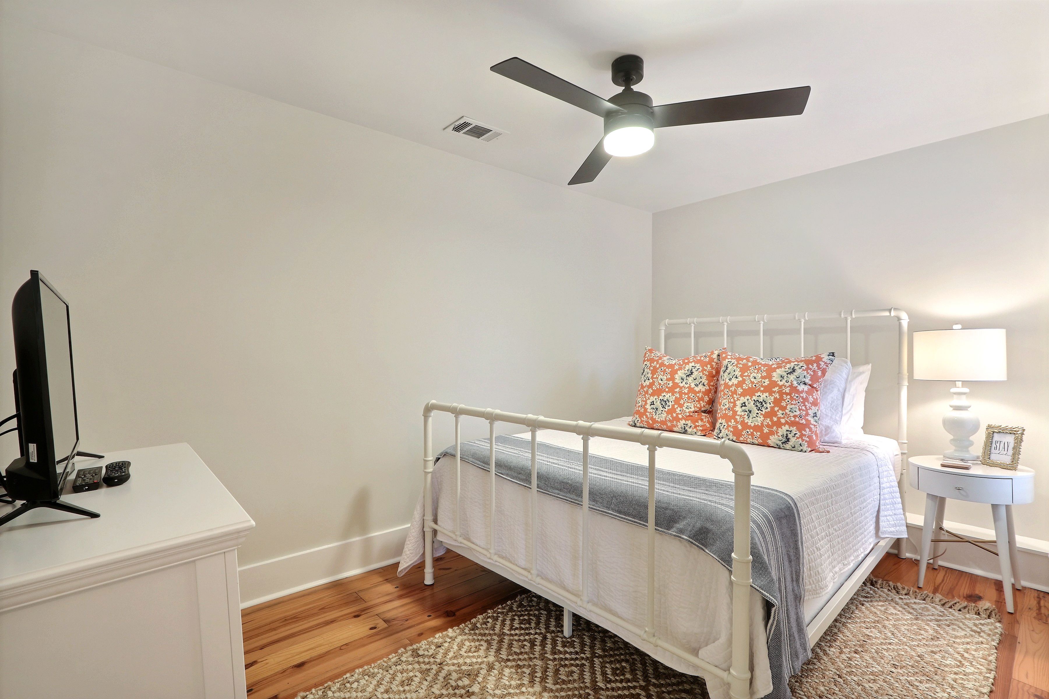 A bedroom with a white bed, a ceiling fan, a bedside table with a lamp, and a TV on a stand.