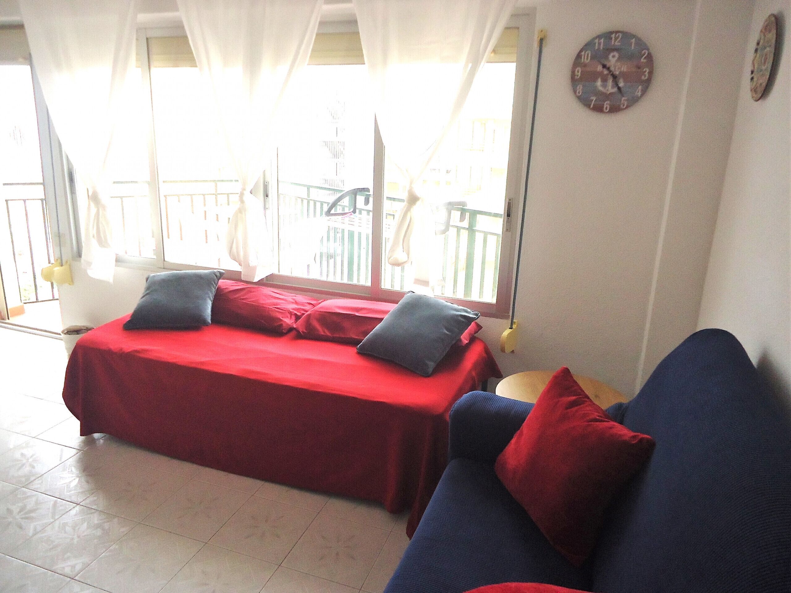A room with a bed, a sofa, a clock, and a balcony with a railing.