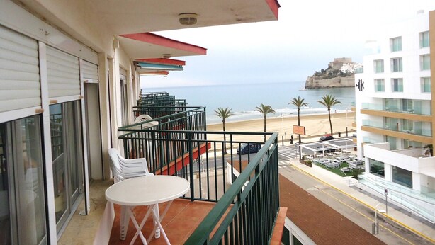 A balcony with a white table and chairs, overlooking a beach and a building with a red roof. - Les Doyes Bl 4 pta 145 (Peñiscola)