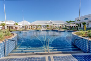 A large outdoor swimming pool with a clear blue water feature, surrounded by palm trees and shaded seating areas.