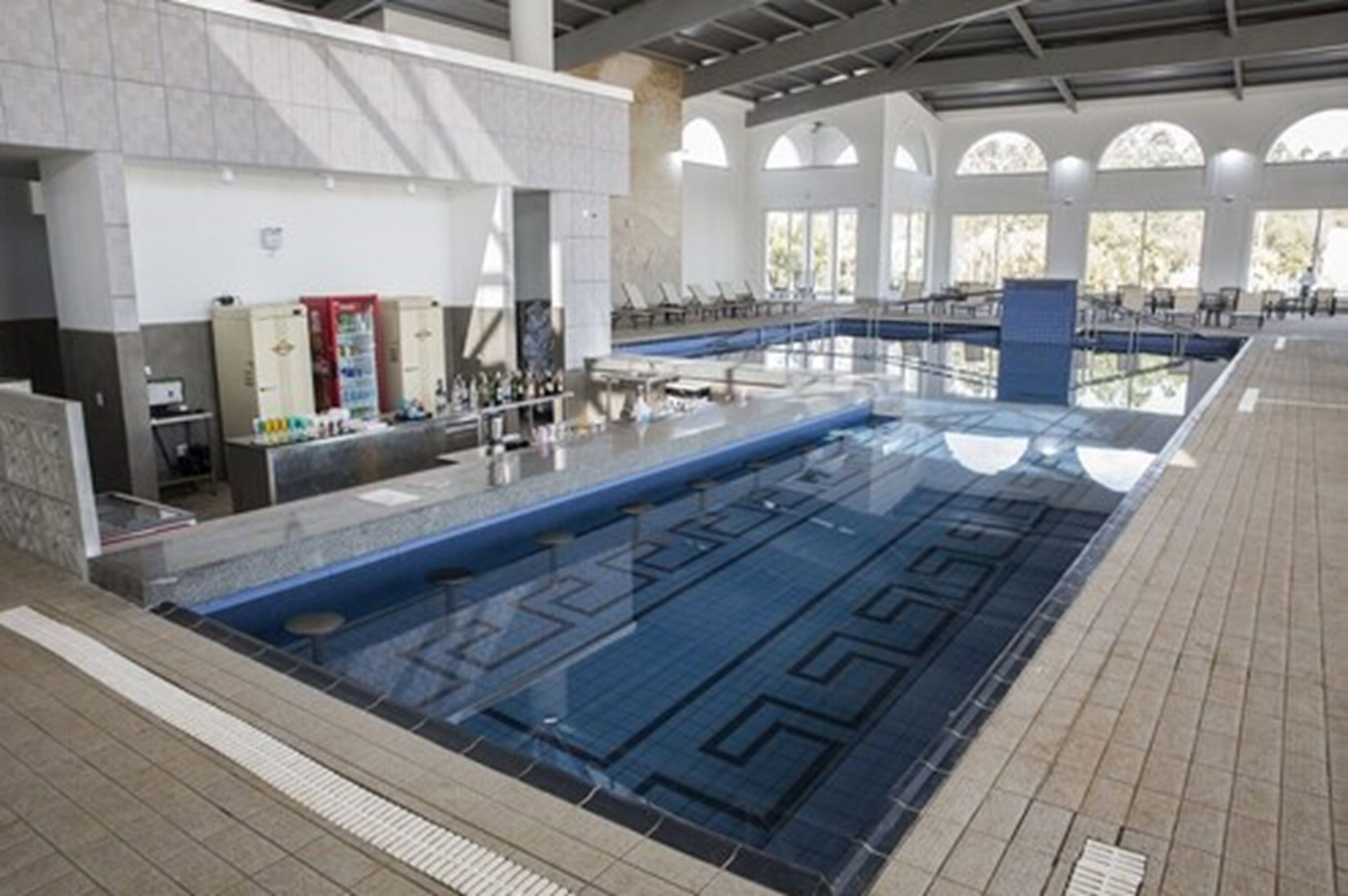 Una amplia piscina interior con suelo de baldosas, zona de vestuarios y mostrador de recepción.