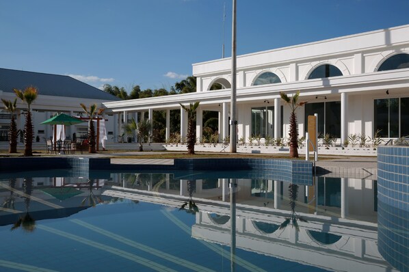 A modern hotel with a large pool, palm trees, and a covered outdoor seating area.