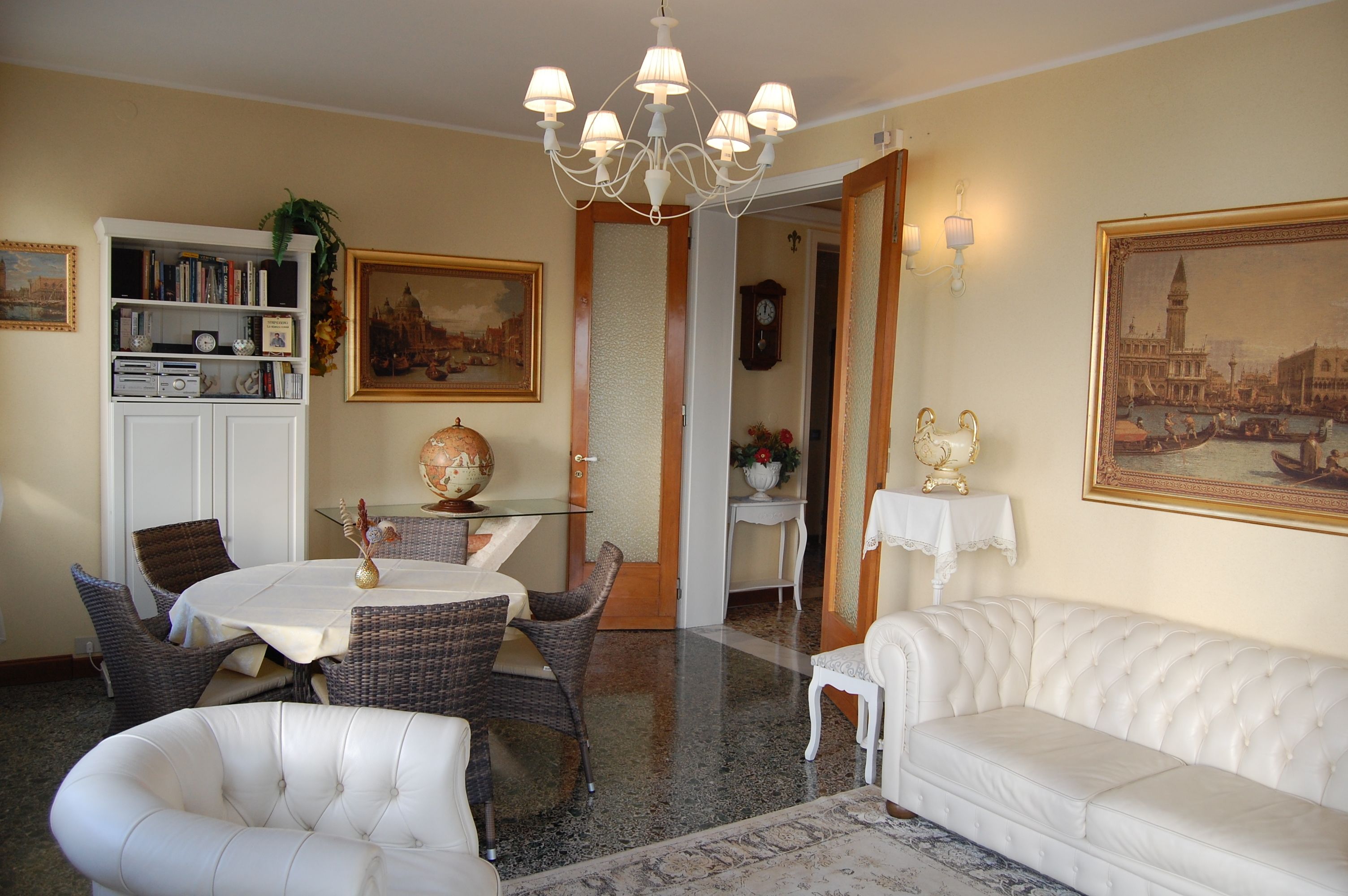 Un coin repas avec des chaises en osier, une table blanche et un lustre. On y trouve également un canapé blanc et une bibliothèque remplie de livres et d’objets décoratifs.