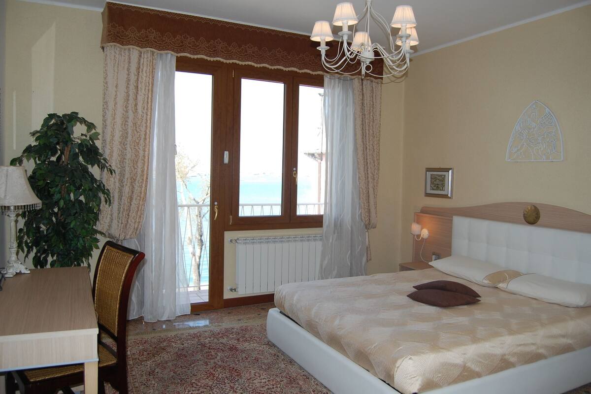 A bedroom with a bed, a desk, a chair, a potted plant, and a view of the sea through a window.