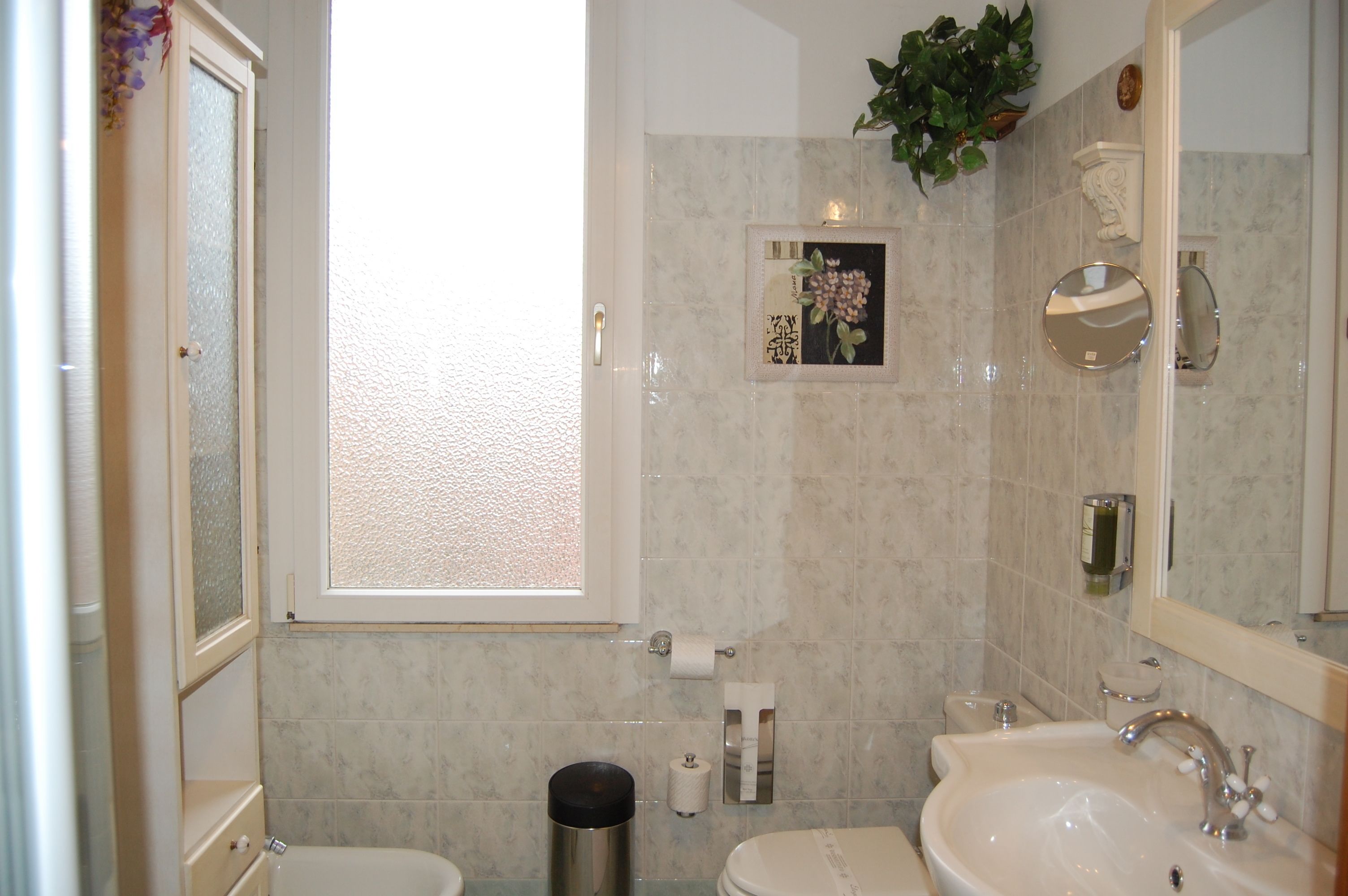 Un baño con inodoro, lavamanos y espejo. Hay una ventana con vistas al exterior.