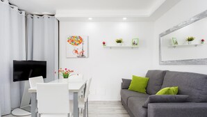 Ein modernes Wohnzimmer mit einem grauen Sofa, einem weißen Esstisch und Stühlen. Ein Flachbildfernseher ist an der Wand montiert, und auf den Regalen stehen dekorative Gegenstände.