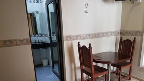 Una habitación con una cama, una mesa de comedor de madera con sillas, un ventanal pequeño y un baño visible a través de una puerta abierta.