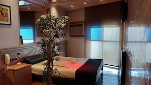A modern hotel room with a large bed, a bedside table, a telephone, a potted plant, and a large window with blinds. - Dallas Hotel (Motel) (Buenos Aires)