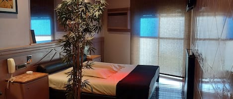 Una habitación de hotel moderna con una cama grande, una mesita de noche, un teléfono, una planta en maceta y un ventanal con persianas.