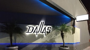 A building facade with the word "DALLAS" illuminated in large letters, two potted palm trees, and a blue-lit ceiling. - Dallas Hotel (Motel) (Buenos Aires)