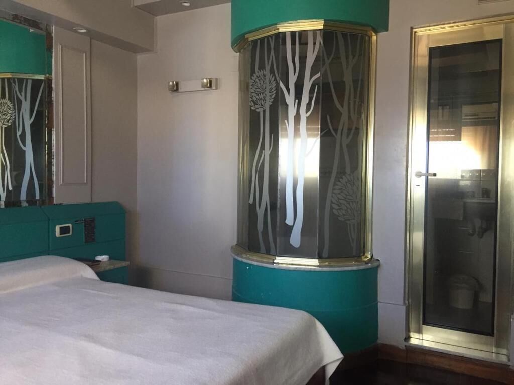 A hotel room with a bed, a teal-colored headboard, a cylindrical decorative piece with tree designs, and a glass door with a reflection of a bathroom.