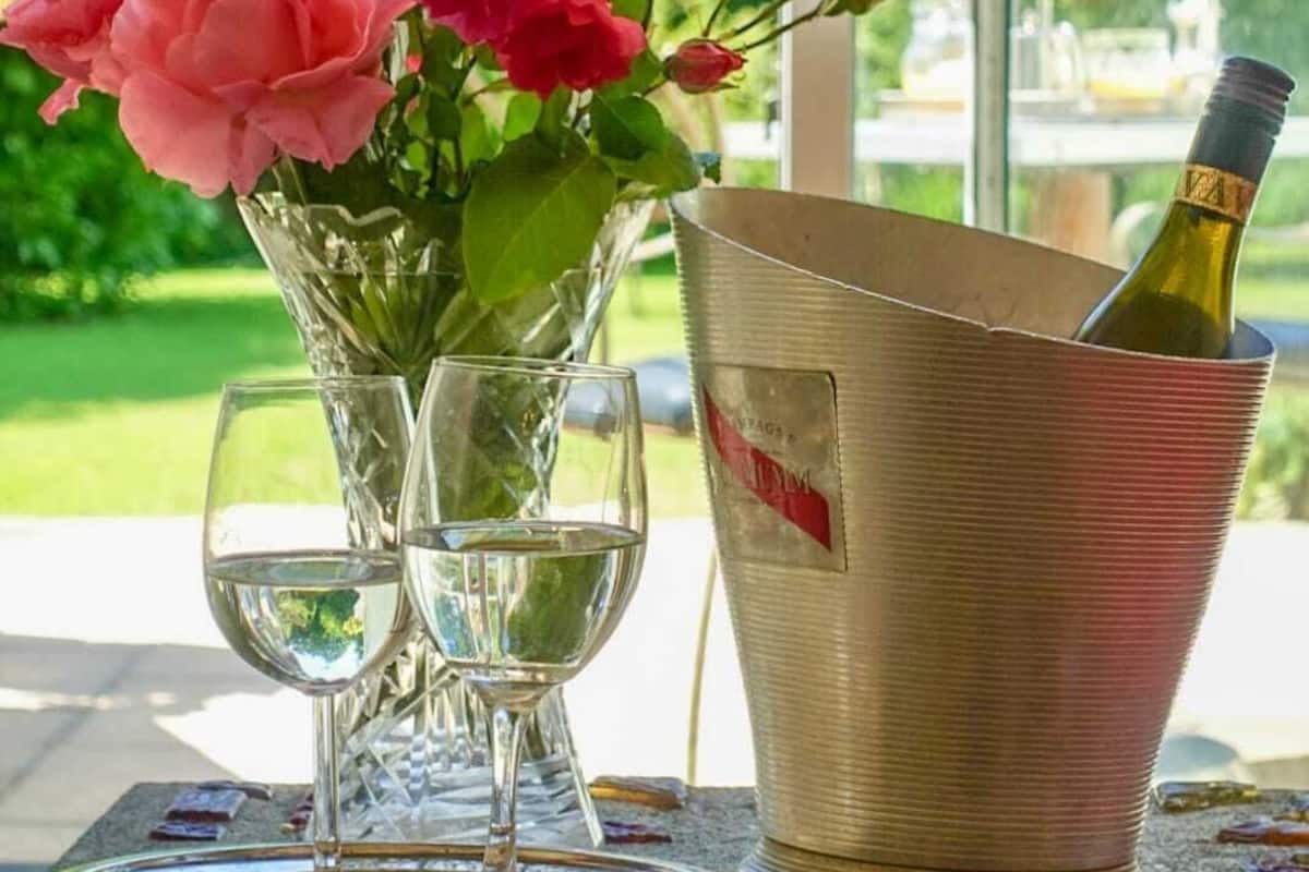 A table with a glass vase of pink roses, two wine glasses, and a metal bucket containing a bottle of wine.