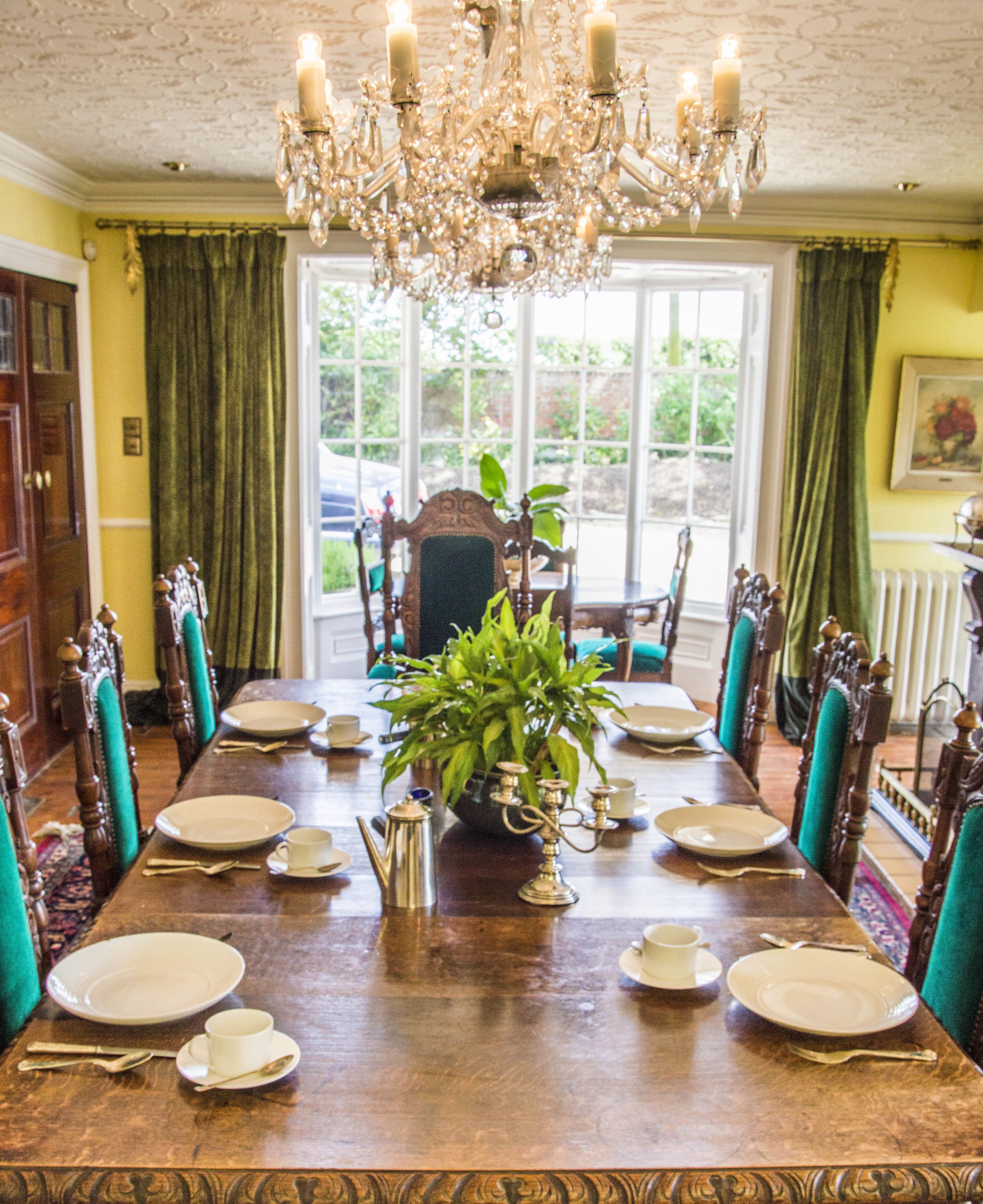 Un comedor con una gran mesa de madera dispuesta para seis personas, rodeada de sillas ornamentadas. Un candelabro cuelga del techo, y una planta en maceta se encuentra en el centro de la mesa.