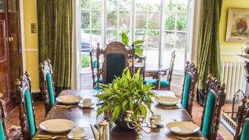 Un comedor con una gran mesa de madera dispuesta para seis personas, rodeada de sillas ornamentadas. Un candelabro cuelga del techo, y una planta en maceta se encuentra en el centro de la mesa.