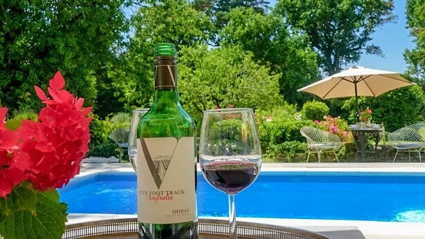 A bottle of wine and a glass of red wine on a tray by a pool with a view of trees and a clear sky. - Cedar House Boutique B&B (Christchurch)