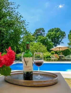 A bottle of wine and a glass of red wine on a tray by a pool with a view of trees and a clear sky. - Cedar House Boutique B&B (Christchurch)