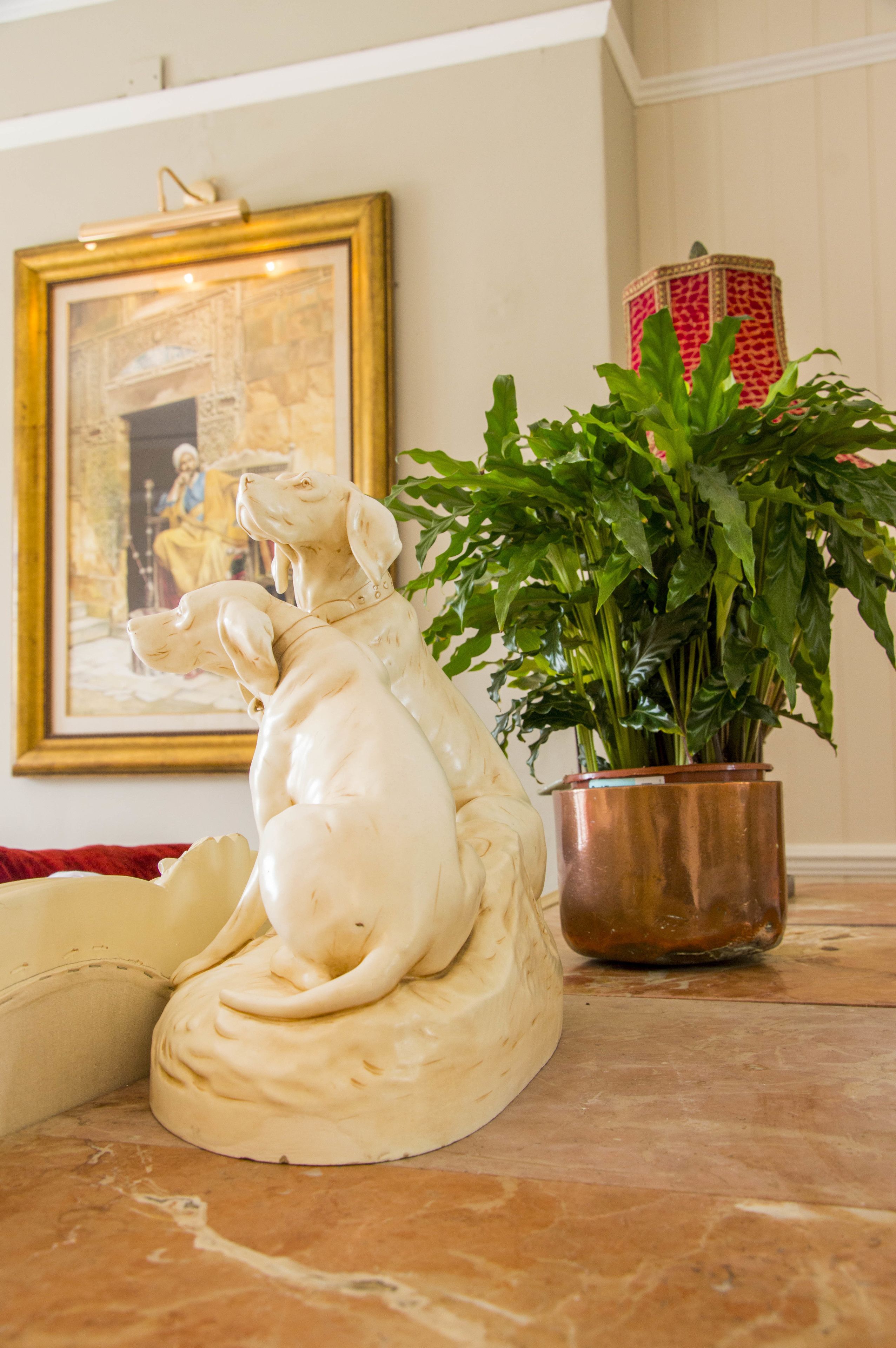 A cream-colored dog sculpture on a tiled floor, a framed painting on the wall, and a potted plant with green leaves.