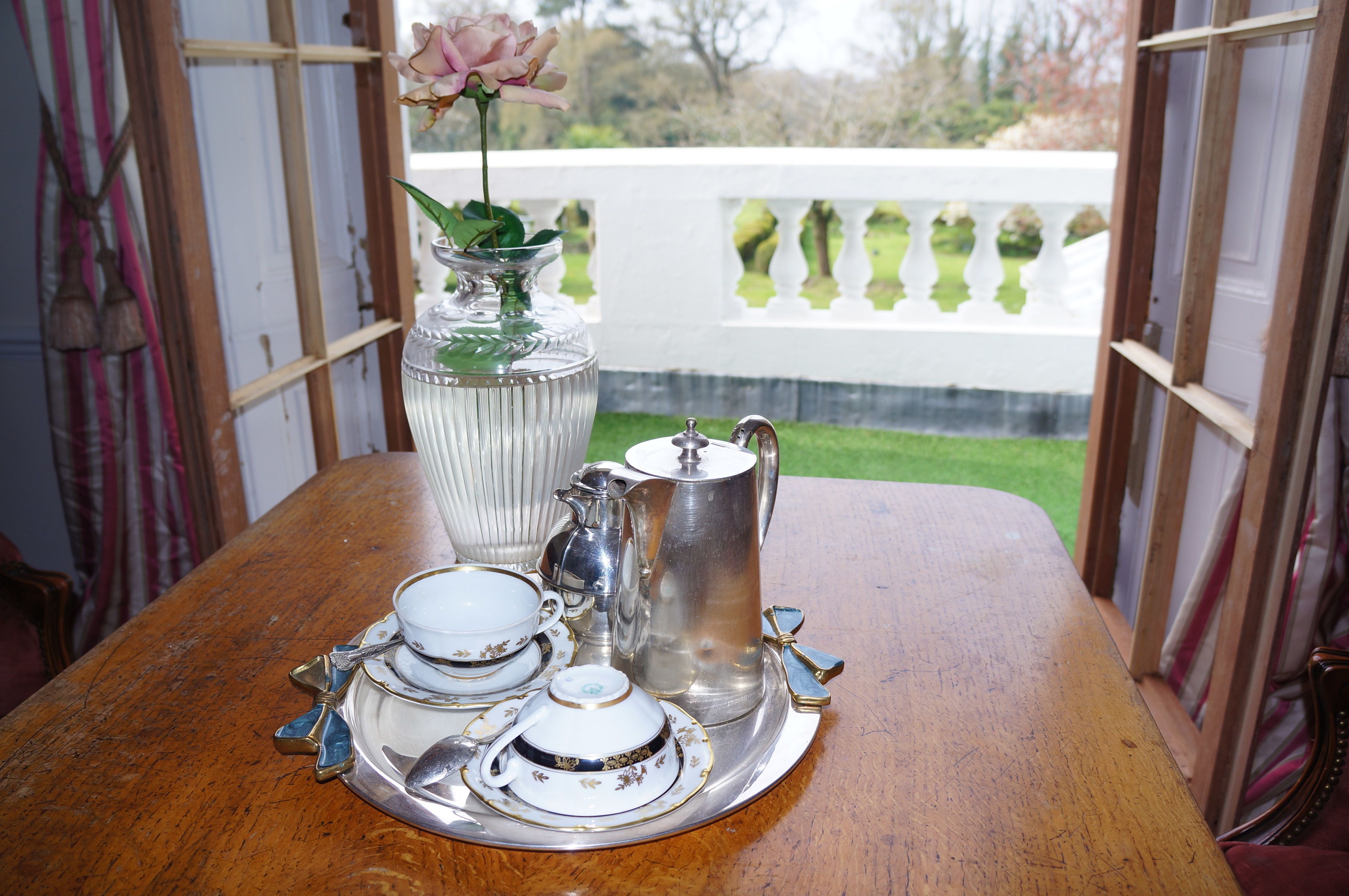 Une table dressée avec un service à thé en argent, comprenant une théière, des tasses et des soucoupes, et un vase contenant une rose.