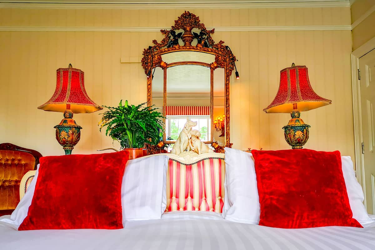 A bed with red and white bedding, a large ornate mirror, two decorative lamps, and a potted plant.