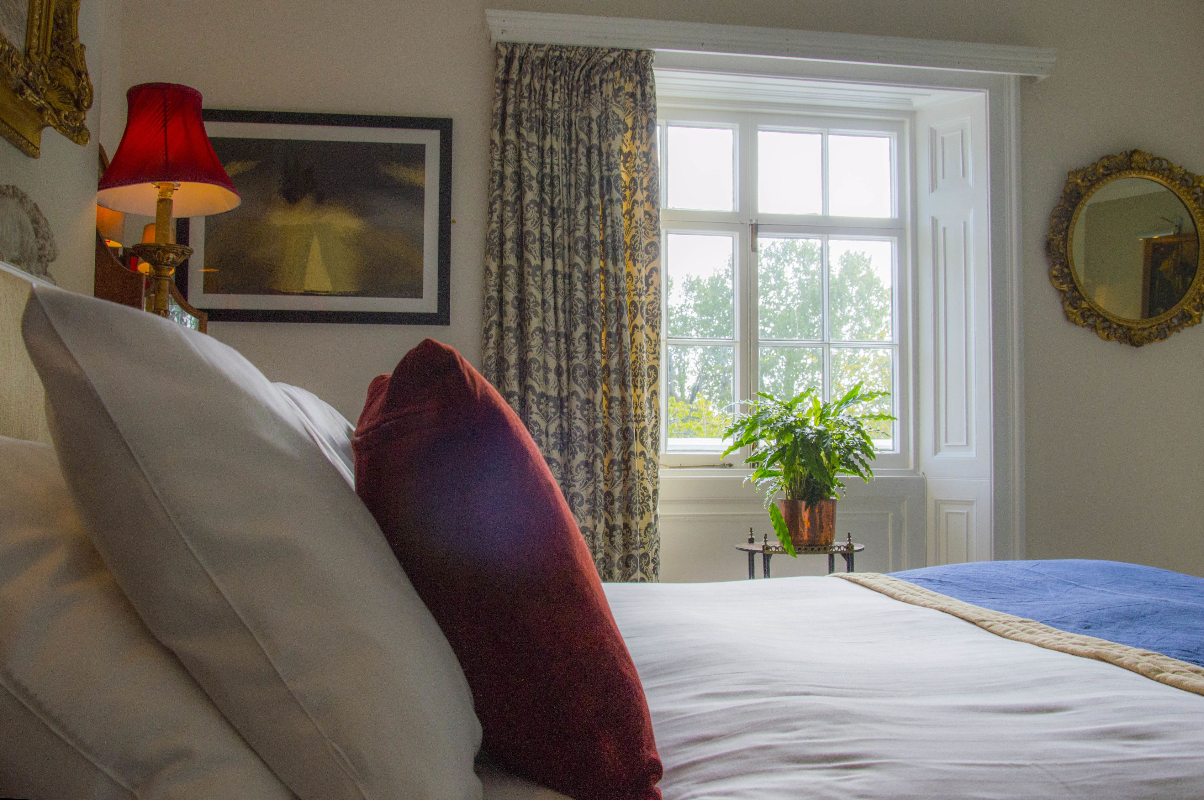 A neatly made bed with white and red pillows, a red lamp, a framed painting, a window with curtains, and a potted plant.