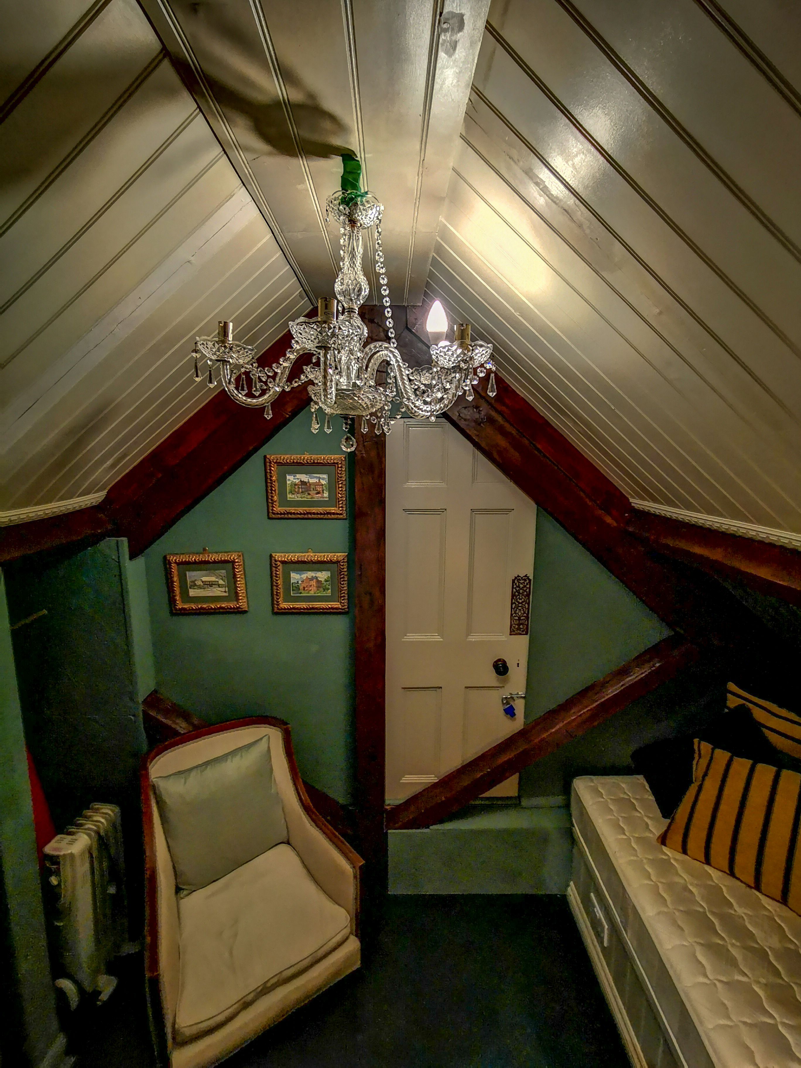 A room with a sloped ceiling, a chandelier, a chair, a bed, and framed pictures on the wall.