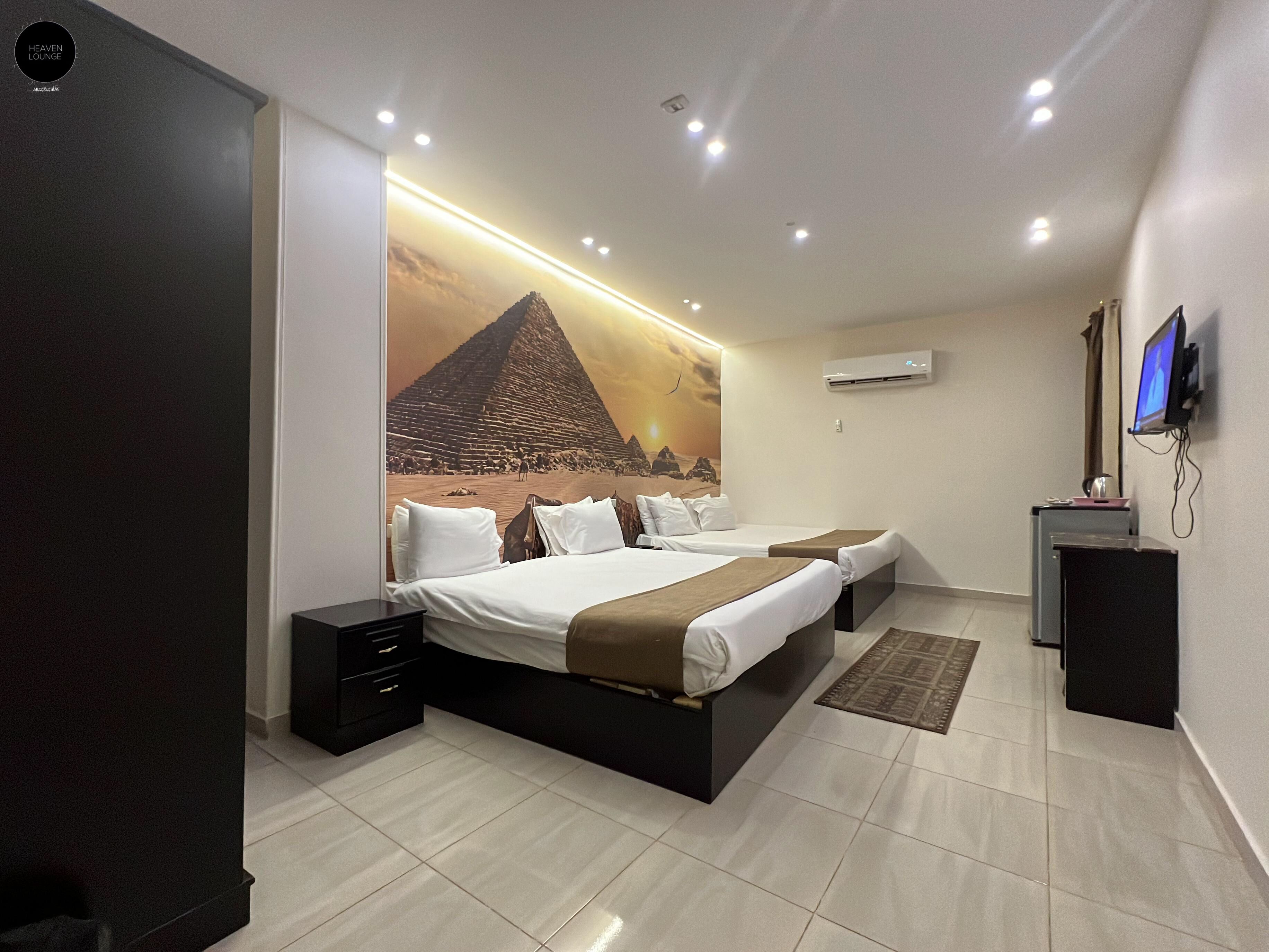 A modern hotel room with a large bed, a desk, a flat-screen TV, and a mural of a pyramid.