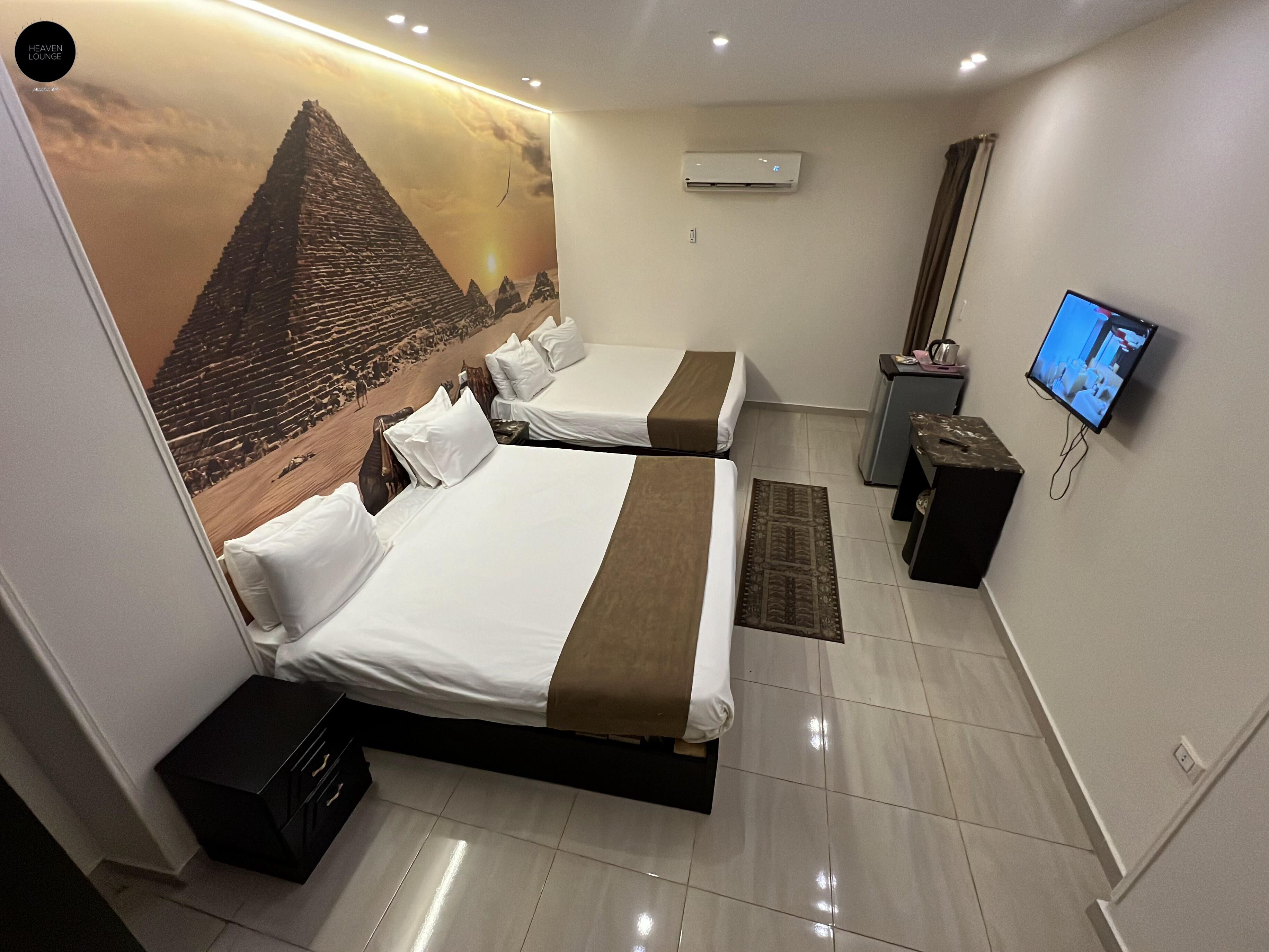 Une chambre d’hôtel avec deux lits, une télévision, un appareil de climatisation et une fresque représentant une pyramide.