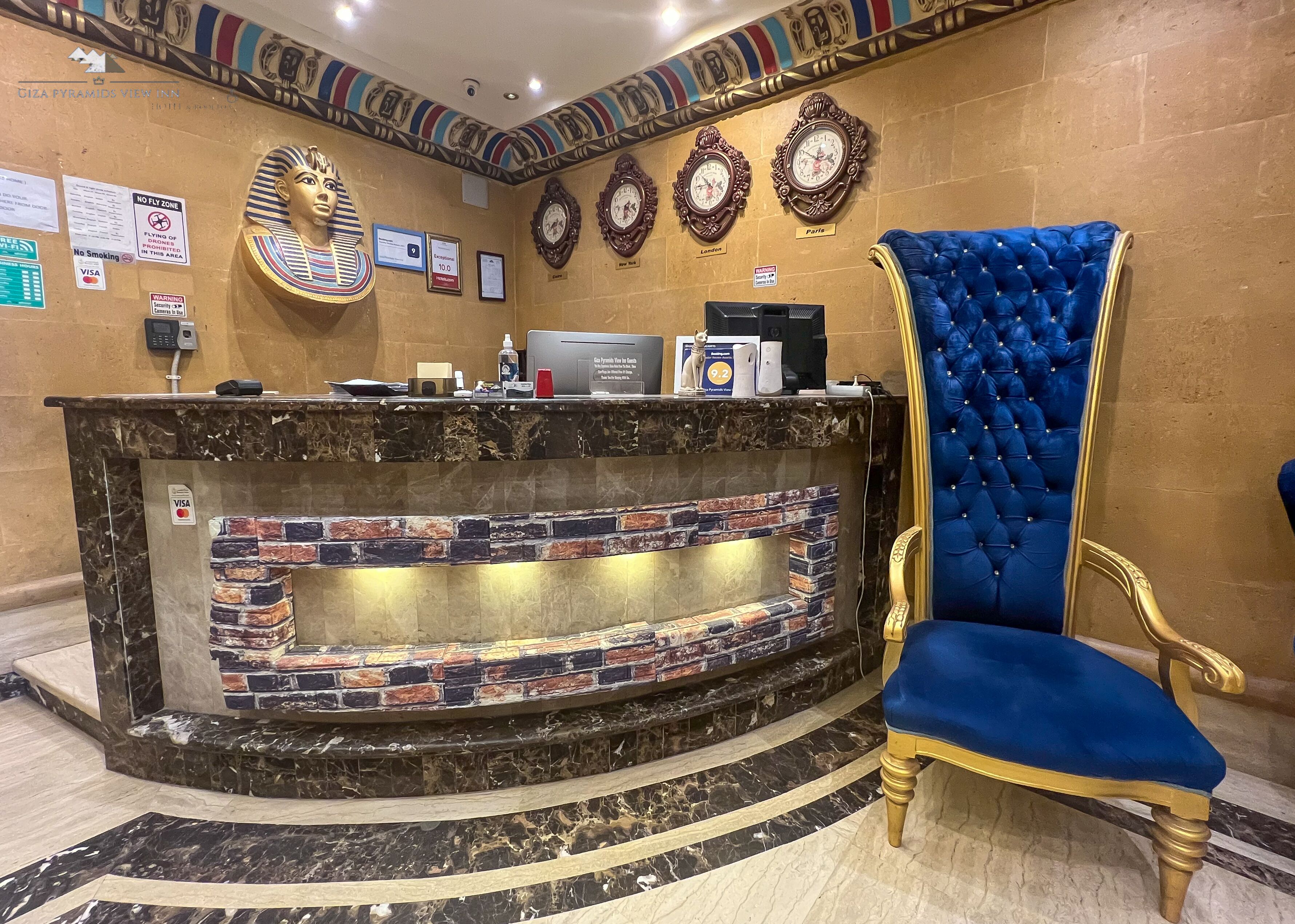 A reception area with a marble counter, a blue upholstered chair, and wall decorations including clocks and a sphinx statue.