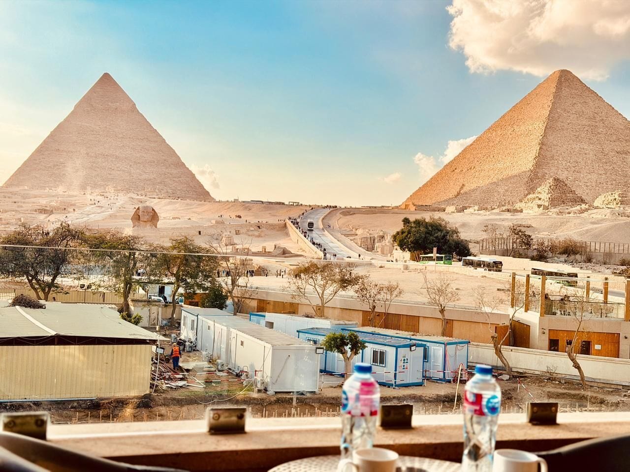 A view of the pyramids with a road leading towards them, a small settlement with buildings, and a table with water bottles and a cup.