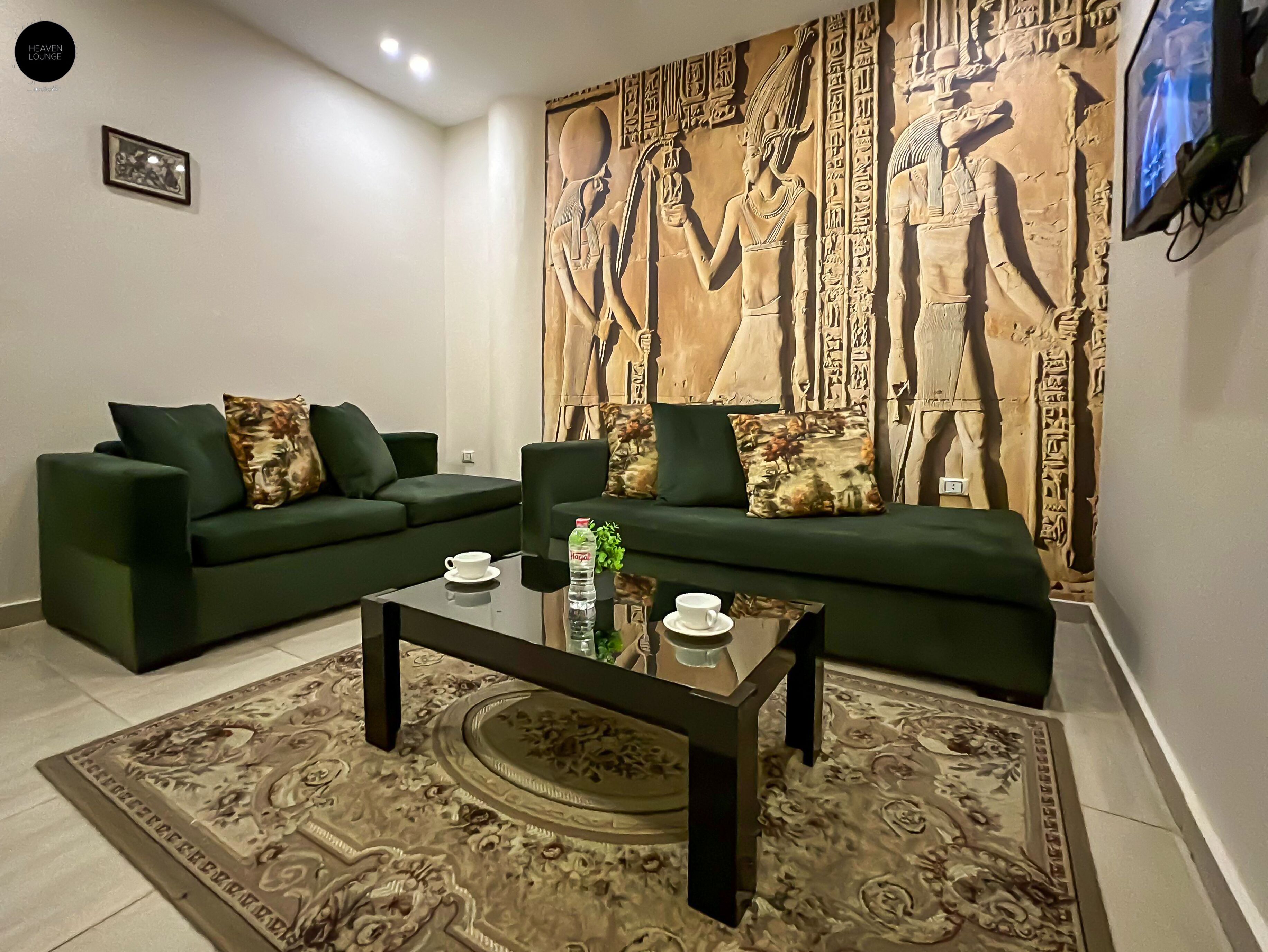 A living room with two green sofas, a coffee table, and a decorative wall with ancient Egyptian reliefs.