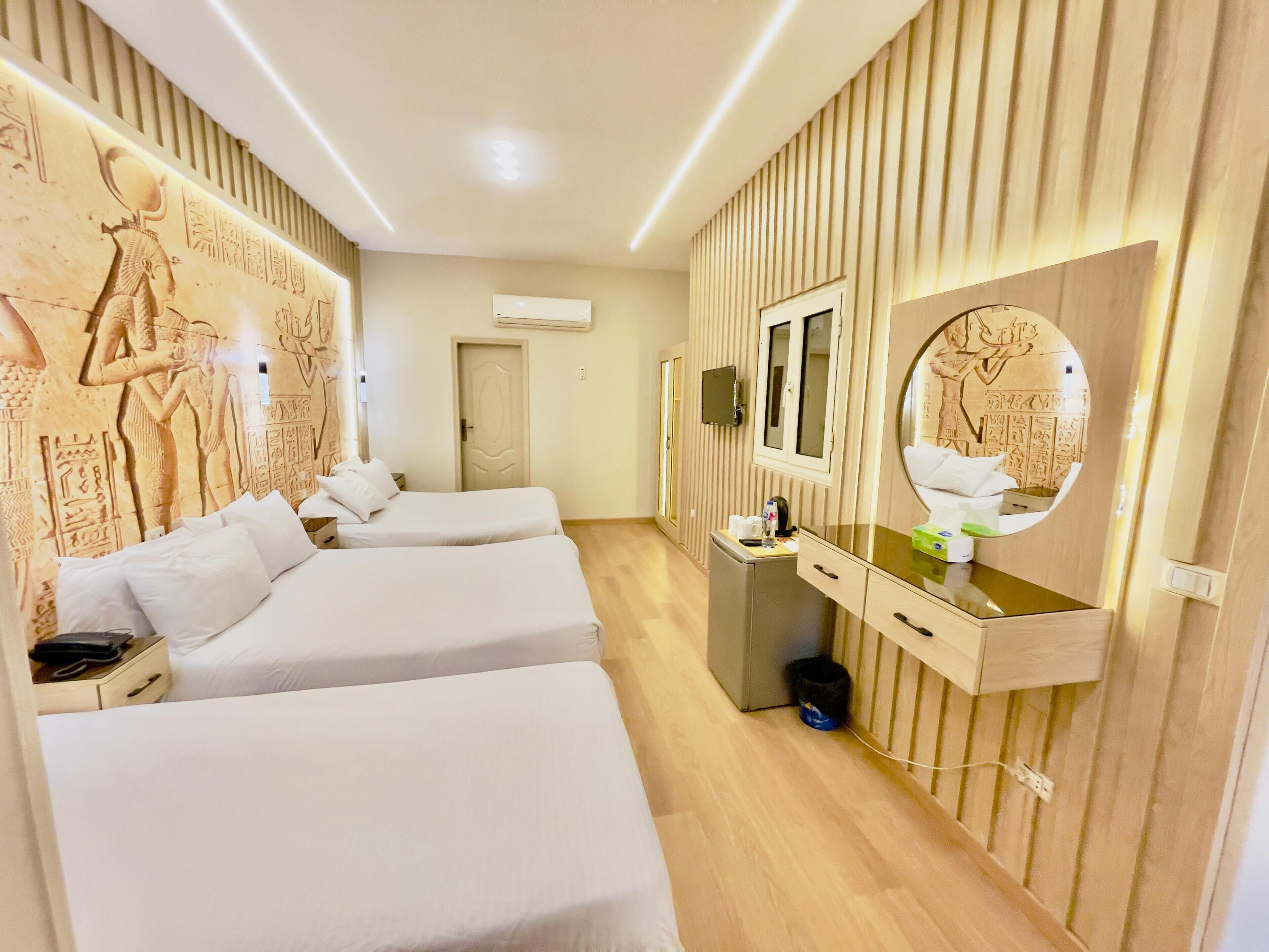 A hotel room with two beds, a minibar, a mirror, and Egyptian wall art.