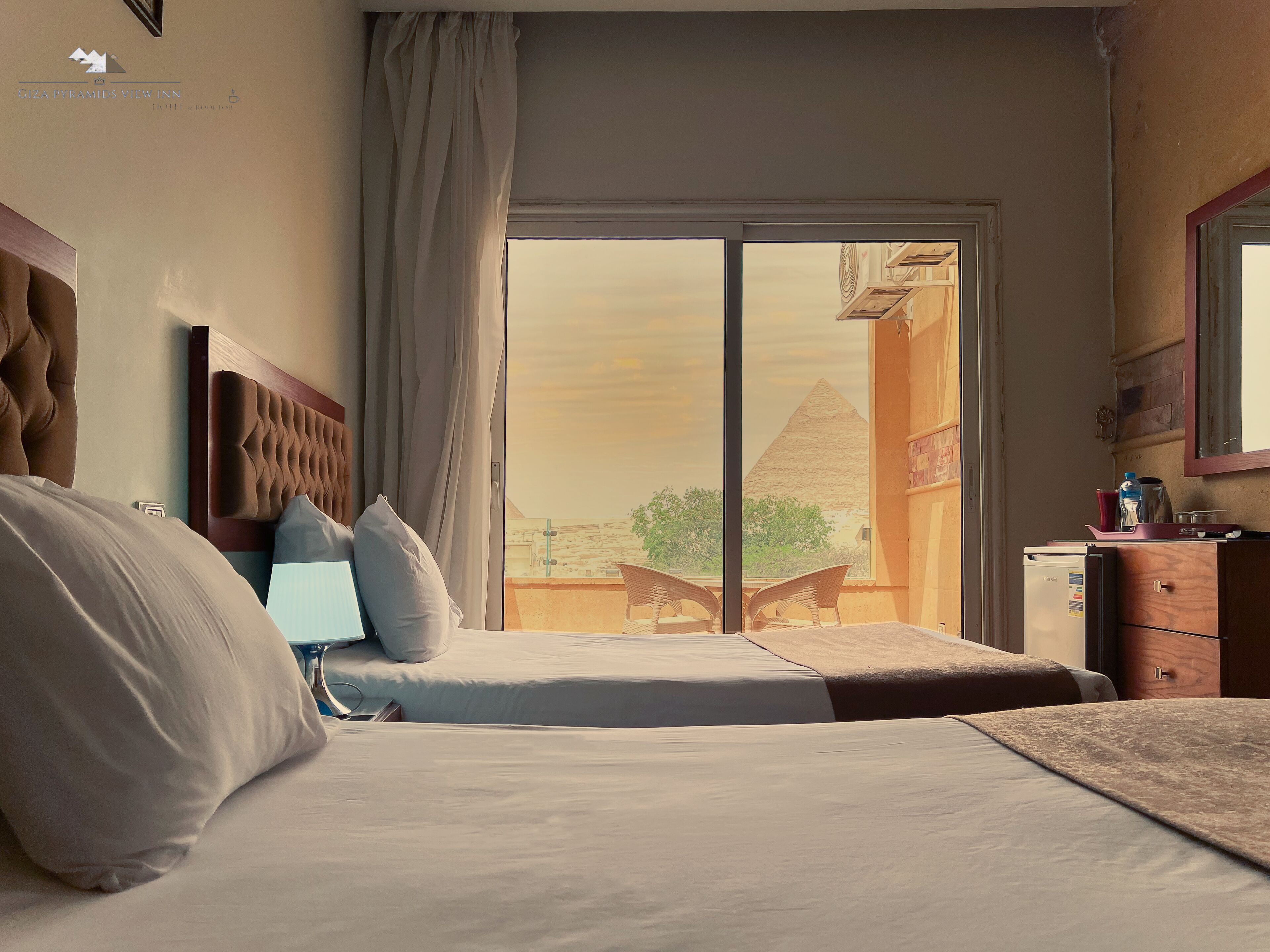 A hotel room with a large bed, a headboard, a nightstand, a dresser, and a view of a pyramid through a glass door.