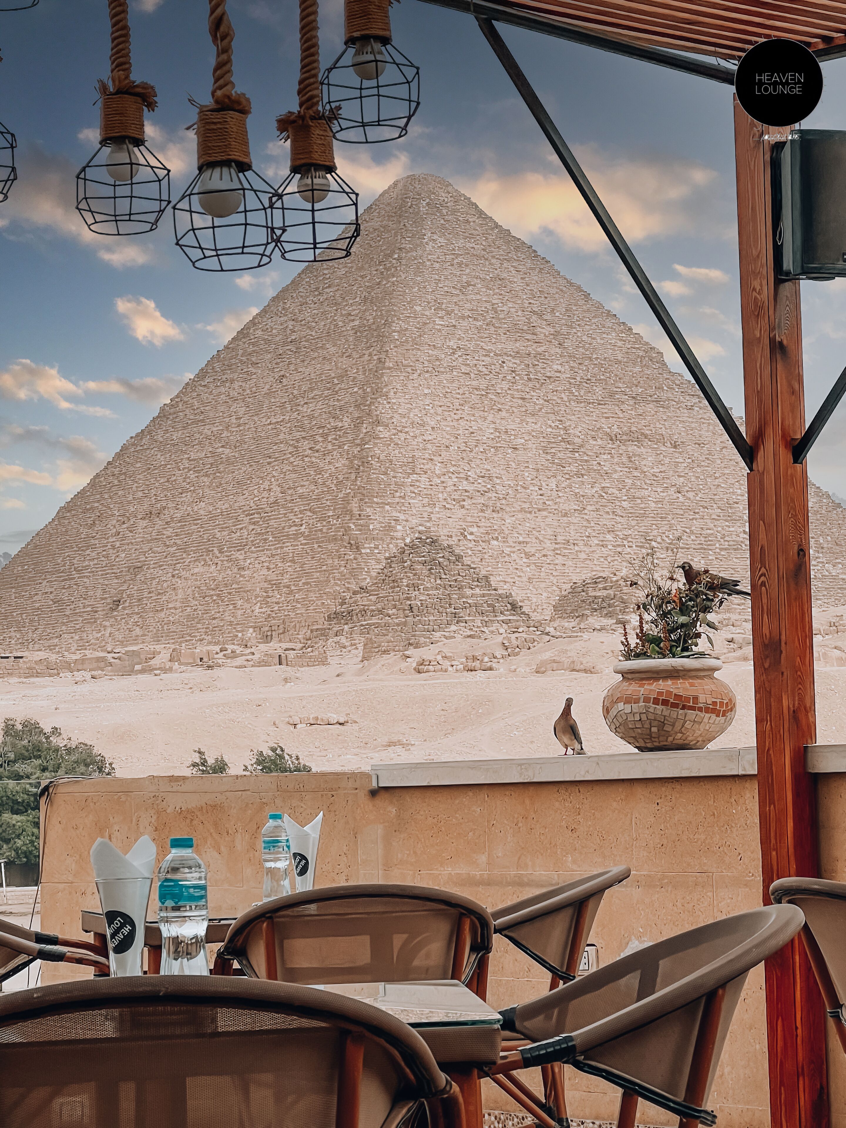 A view of a pyramid from a covered outdoor seating area with wicker chairs and hanging light fixtures.