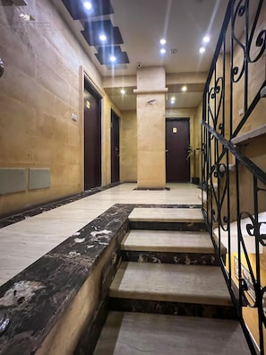 A hotel corridor with marble flooring, a central staircase, and a metal railing.