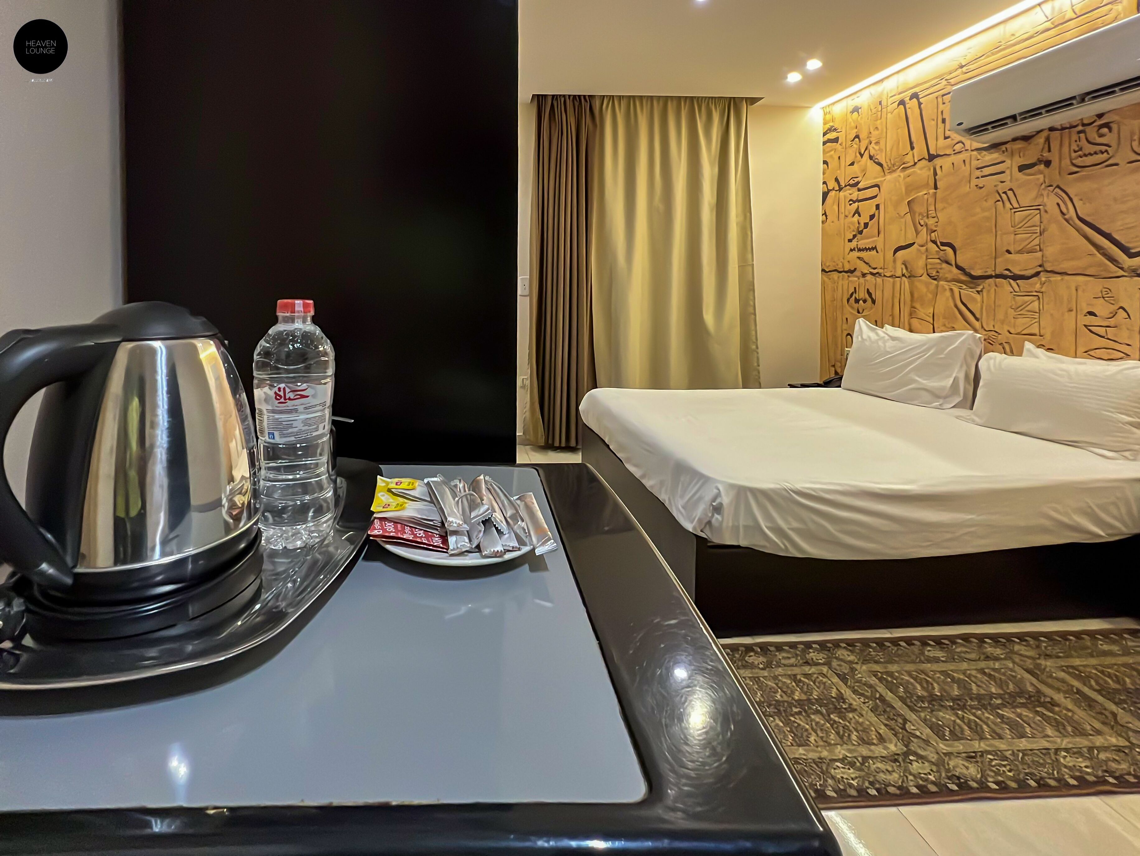 A hotel room with a bed, a bedside table with a kettle, a bottle of water, and some snacks.
