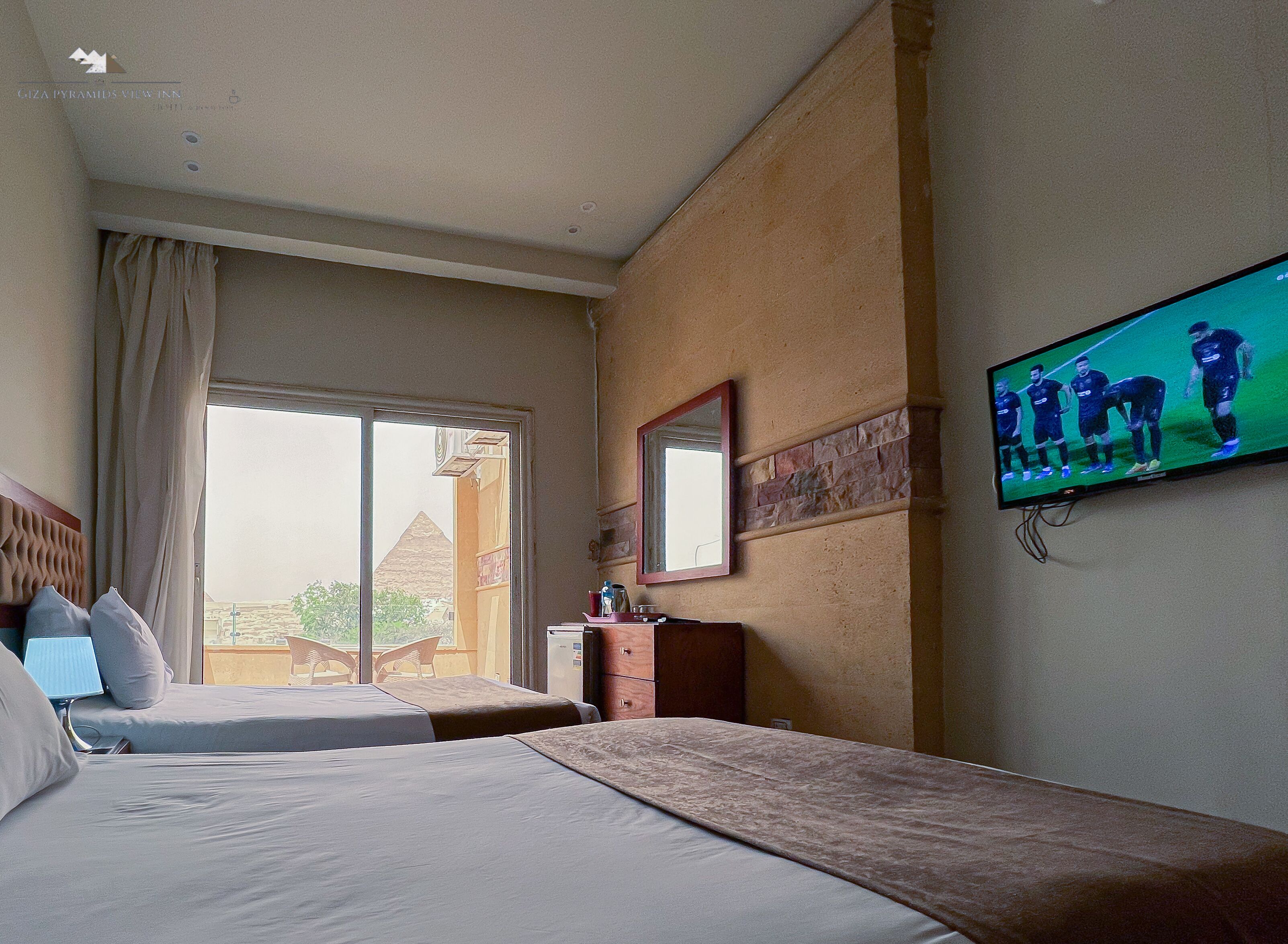 A hotel room with two beds, a TV mounted on the wall, a window with curtains, and a view of a pyramid outside.